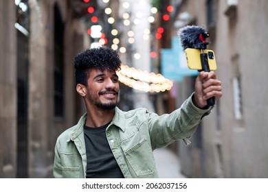 African-American influencer recording on the street with his mobil camera and professional microphone while broadcasting live to his followers with a natural expression and smiling. Bloggers concept.