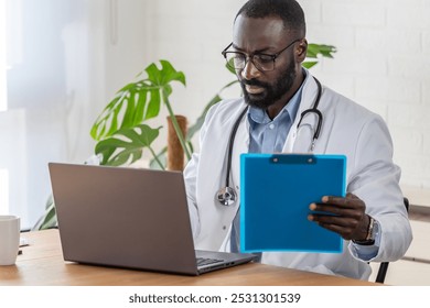 An African-American male doctor in a white coat and glasses is working on a tablet in a modern office. He is focused and engaged in his task, possibly conducting research or reviewing patient records. - Powered by Shutterstock