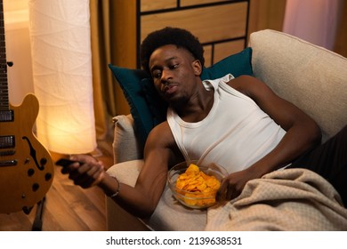 African-American Male Muscular Athletic Relaxes In Evening On Couch Watching TV And Eating Chips From Bowl, Student Switches Channels, Programs With Remote Control