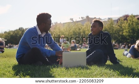 African-American male professor discussing exam results with student sitting on lawn near school. Portrait of teacher and teenage boy sitting on grass on schoolyard and talking with laptop