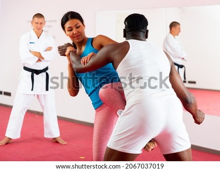 African-american man and Asian woman exercising fighting moves together in gym, their trainer in kimono standing and watching.