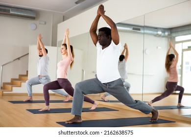 African-american man exercising warrior one pose during group yoga training in gym.