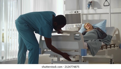 African-American Man Nurse Adjusting Bed For Woman Patient. Young Sick Female Resting In Hospital Bed While African Male Doctor Lifting It Up In Clinic Room