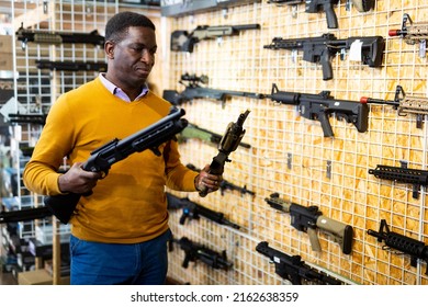 African-american Man Standing In Salesroom Of Gun Shop And Holding Shotgun And Assault Rifle In Hands.