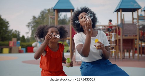 African-American mother and daughter play soap bubbles at playground. Happy woman and preschool girl sitting on bench and blowing soap bubbles together - Powered by Shutterstock