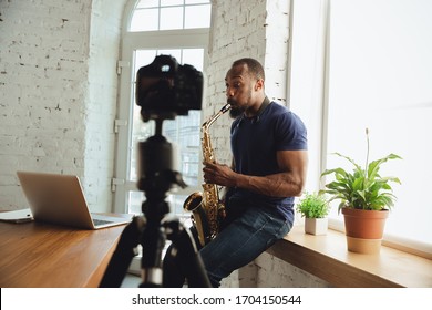 African-american musician playing saxophone during online concert at home isolated and quarantined. Using camera, laptop, streaming, recording courses. Concept of art, support, music, hobby, education