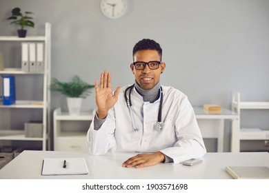 African-American Online Doctor In Lab Coat Sitting At Desk In His Office And Waving Hand At Camera, Welcoming Clients On Medical Video Channel Or Saying Hello Or Goodbye To Patient During Video Call