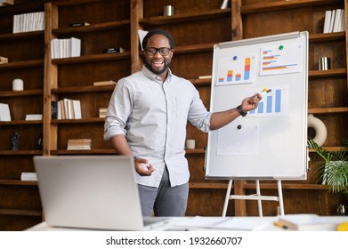 An African-American online teacher, tutor, business coach is holding webinar, points at whiteboard with graphs, explains to online audience on the laptop, biracial man makes a presentation online