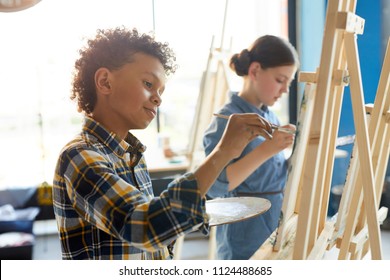 African-american schoolboy with palette and paintbrush painting picture on easel during individual work - Powered by Shutterstock