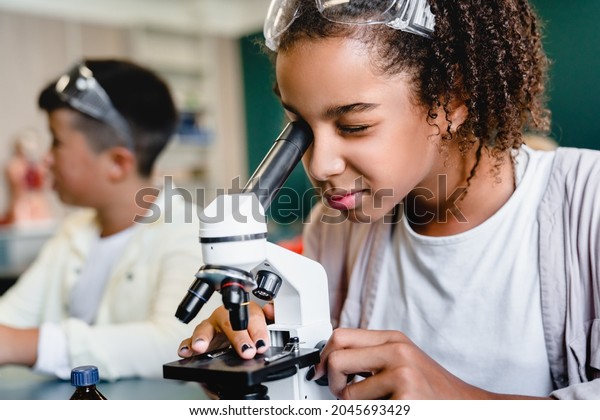 African-american schoolgirl pupil student using\
working with microscope at biology chemistry lesson class at school\
lab. Science lesson\
concept