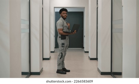 African-American security cameras installer in uniform stands in corridor with laptop, looks at camera. He sets up CCTV cameras in office using computer software. Surveillance system concept. - Powered by Shutterstock