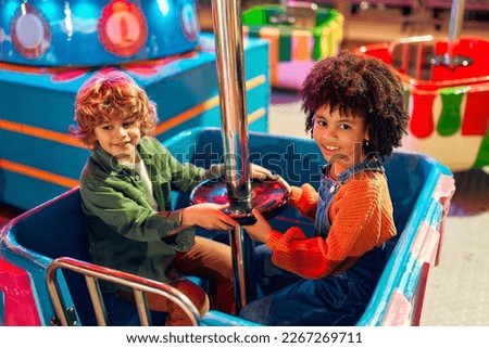 An African-American sweet girl child with afro curls and a cute Caucasian boy ride together on a carousel in an amusement park in the evening on weekend and have fun.