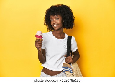 African-American teen girl with summer look, beach bag, enjoying ice cream on yellow backdrop. - Powered by Shutterstock