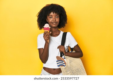 African-American teen girl with summer look, beach bag, enjoying ice cream on yellow backdrop. - Powered by Shutterstock