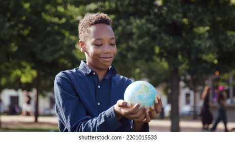 African-American teenage boy looking at globe of the earth standing outdoors. Schoolboy smile holding globe outside school campus. Nature and ecology concept