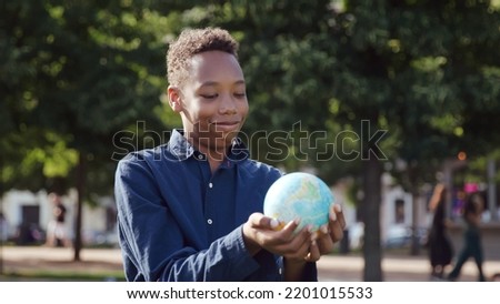 African-American teenage boy looking at globe of the earth standing outdoors. Schoolboy smile holding globe outside school campus. Nature and ecology concept