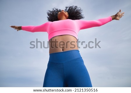 African-American woman with afro hair and sportswear, with fluorescent pink t-shirt and leggings, jumping into the sky, seen from below. Fitness concept, sport, street, urban, happiness.