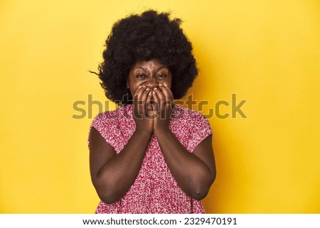 African-American woman with afro, studio yellow background laughing about something, covering mouth with hands.