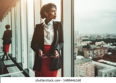 An African-American woman entrepreneur a digital tablet is leaning against a panoramic window of an office business skyscraper and looking in a distance; biracial businesswoman with a tablet pc