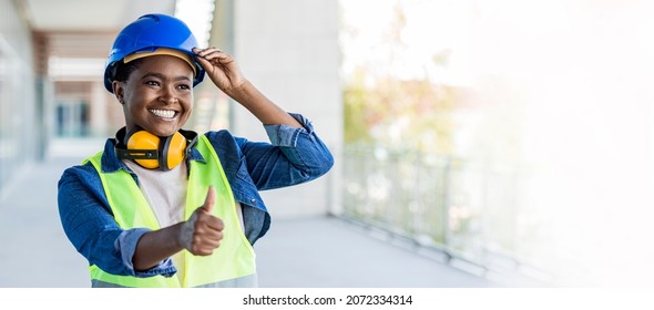 A African-American woman in her 30s working at a construction site, wearing a hardhat, safety goggles and reflective vest. She is looking over the camera with a confident expression, showing thumb up.