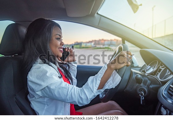 African-American woman inside\
a car with a mobile phone in her hand and the other hand on the\
steering wheel
