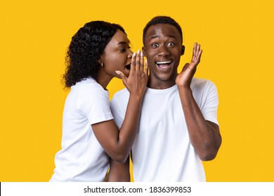African-american woman sharing secret or whispering gossips into her boyfriend's ear, yellow studio background. Black lady whispering words of love to her amazed man who screaming and gesturing