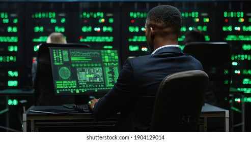 African-American IT worker sitting in server room behind workplace working with database on computer. System software concept. View from back.