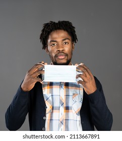 African-american Young Man Taking Off Or Putting On Medical Mask. No More Covid Concept. Not Happy Or Tired Young Man Taking Off Medical Mask. African Man With Medical Mask In Hands. Isolated On Grey.