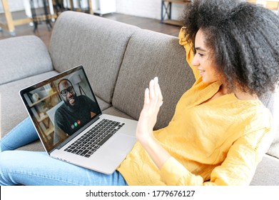 African-American Young Woman Is Using App On Laptop For Video Meeting With Friend Or Colleague. Biracial Woman With A Laptop Lays On The Couch And Waving Hello To Black Guy On Video Screen