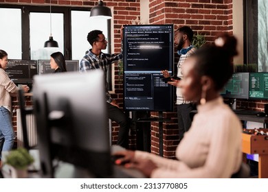 Africann american system engineers analyzing source code looking for errors, checking big tv monitor in it agency office. Software developers working on cyber security innovation in it agency.