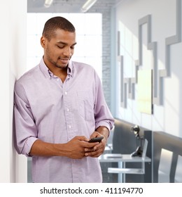Afro American Casual Office Worker Using Mobile Phone, Checking Out Business. Smiling, Leaning Against Wall.