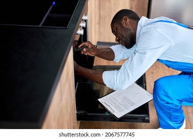Afro american confident repairman in uniform working, fixing broken oven in kitchen using tools. Black guy in blue workwear is repairing. Repair service concept. Side view portrait - Shutterstock ID 2067577745