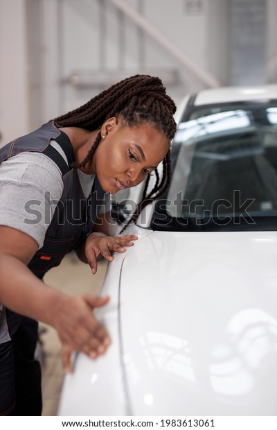 Afro american Female auto\
mechanic touching surface of white repainted and clean car body in\
auto repair shop. Pretty black woman Mechanic in uniform working in\
workshop