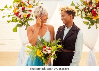 Afro American Female In The Role Of The Groom And Short Blonde Haired Bride In Blue Dress In Ceremony Under Wedding Arch In Tropical Beach
