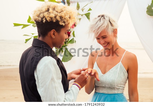 afro american female in the role of the groom and\
short blonde haired bride in blue dress in ceremony under wedding\
arch in tropical beach
