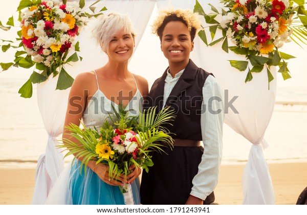 afro american female in the role of the groom and\
short blonde haired bride in blue dress in ceremony under wedding\
arch in tropical beach