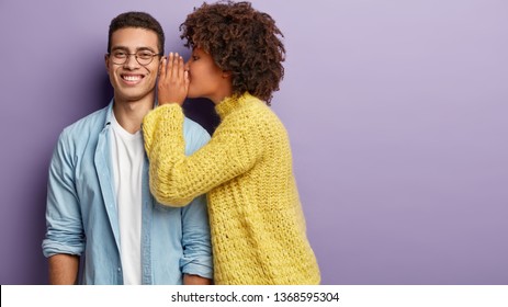 Afro American female whispers secret to boyfriend who has cheerful expression, gossip together, wear casual clothes, stands against purple background with blank space. Diverse couple indoor.