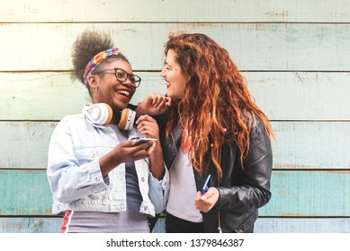 Afro American And Latin American Girls Using Mobile Phone Outdoors. Latin American Teenager Having Fun. Two Girls Using Cellphone And Laughing.Youth Concept.