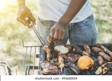 Afro American Man Cooking Meat On Barbecue - Chef Putting Some Sausages And Pepperoni On Grill In Park Outdoor - Concept Of Eating Outdoor During Summer Time - Vintage Retro Filter With Sun Halo Flare