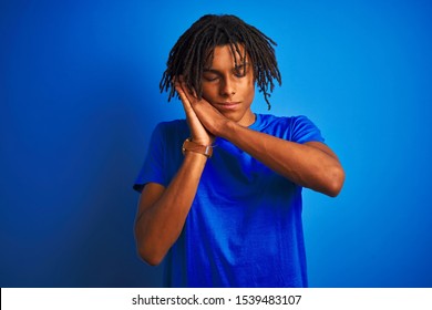 Afro american man with dreadlocks wearing t-shirt standing over isolated blue background sleeping tired dreaming and posing with hands together while smiling with closed eyes.