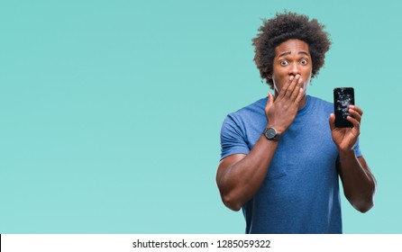 Afro american man holding broken smartphone over isolated background cover mouth with hand shocked with shame for mistake, expression of fear, scared in silence, secret concept