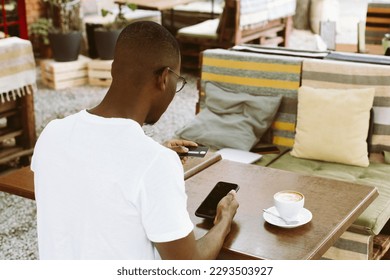 Afro american man sitting at table in cafe with cup of coffee on summer veranda, holding smartphone and entering number of credit card back view. Money, cashless payments, online shopping payment. - Powered by Shutterstock