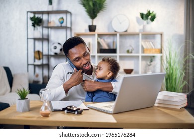Afro american man talking on mobile, working on laptop and carrying baby son. Male freelancer sitting at home office and using modern gadgets.