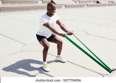 Afro American Man In White T-shirt And Black Shorts Doing Exercise On His Hands Squatting With Elastic Fitness Tape Outside