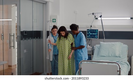 Afro American Nurse With Doctor Helping Patient To Stand Up From Bed Holding Intravenous IV Fluid Drip Bag After Medical Surgery In Hospital Ward. Practitioner Monitoring Sickness Recovery