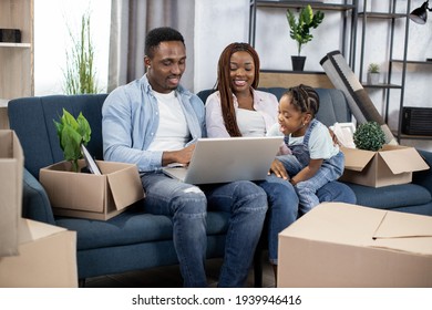 Afro american parents sitting on couch with their cute daughter and using laptop. Young family taking break during removing to new flat.