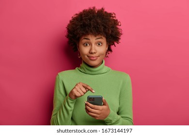 Afro American teenager points at modern smartphone display, recommends download new application, advertises new game, received notification in online chat, wears casual green jumper, poses indoor - Shutterstock ID 1673144197