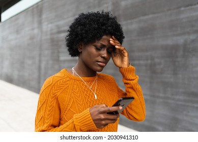 Afro american woman with an orange sweater worried looking at her phone outside in the city. - Powered by Shutterstock