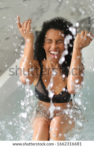 Afro american young smiling woman in black swimsuit taking bath and playing with splashing water. Concept of bathroom photo shoot and morning hygiene.
