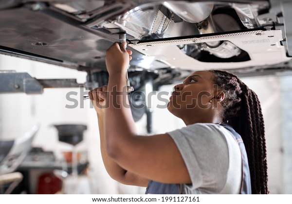 Afro auto mechanic work in garage, car service\
technician woman in overalls check and repair customer car at\
automobile service center, inspecting car under body and suspension\
system